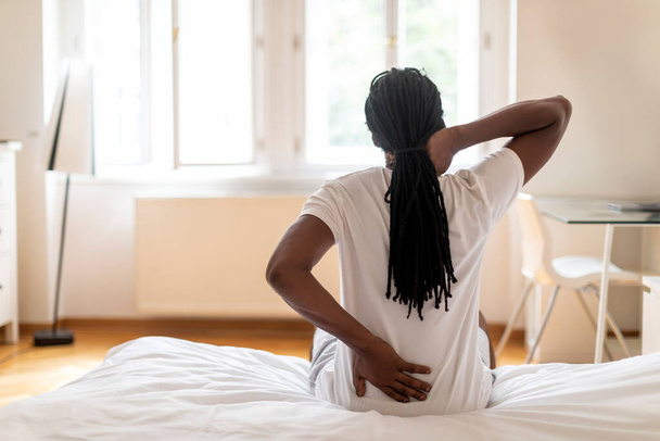 Nierozpoznawalny czarny facet z dredami cierpiący na ból szyi i pleców podczas budzenia się rano, Afroamerykanin mający plecy, siedzący na łóżku w domu i pocierający strefy pocierania, widok z tyłu - Zdjęcie, obraz