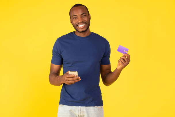 joyeux noir acheteur masculin achats à l'aide de téléphone cellulaire et carte de crédit posant sourire à la caméra en studio sur fond jaune. Mobile Banking App Concept - Photo, image
