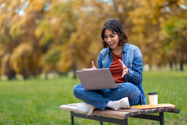 Ekscytowana młoda arabska studentka korzystająca z laptopa podczas siedzenia na ławce w parku miejskim, Millennial Middle Eastern Woman Emocjonalnie reagująca na dobre wiadomości online, z podnieceniem - Zdjęcie, obraz