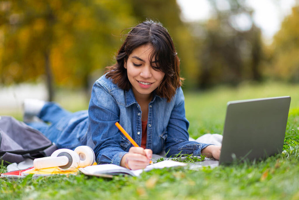 Uzaktan çalışma. Diz üstü bilgisayarlı genç bir kadın dışarıda sınava hazırlanıyor, gülümseyen Orta Doğulu bir kadın çevrimiçi derse katılıyor, bilgisayar kullanıyor ve çimlerde yatarken notlar alıyor. - Fotoğraf, Görsel