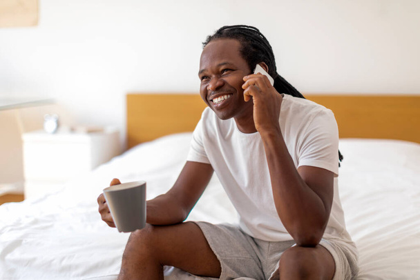 自宅で朝。ベッドルームでベッドに座っている間、陽気な黒人男性が携帯電話で話してコーヒーを飲む、アフリカ系アメリカ人の男が熱い飲み物を持っていて、快適な電話を楽しんで、無料のスペース - 写真・画像