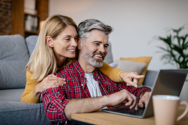 Cheerful mature european wife hugging husband, look at pc, playing online game in cozy living room interior. Entertainment with device, online shopping, social media chat during covid-19 pandemic - Photo, image