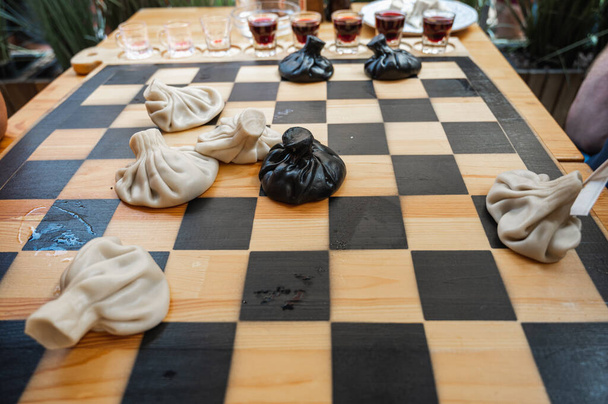 Prato tradicional georgiano, khinkali Kalakuri, em mesa de madeira, tabuleiro de xadrez, rústico, vista lateral, massa preta e leve, jogando xadrez. Foco seletivo, visão lateral. Pratos de carne e queijo - Foto, Imagem