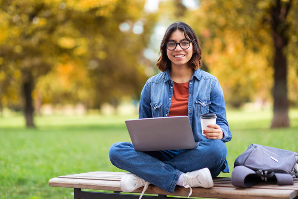 Usmívající se mladá žena z Blízkého východu pomocí notebooku a pití kávy venku, veselý Arab Ženské Student Příprava na zkoušku, zatímco sedí na lavičce v parku města, těší dálkové vzdělávání - Fotografie, Obrázek