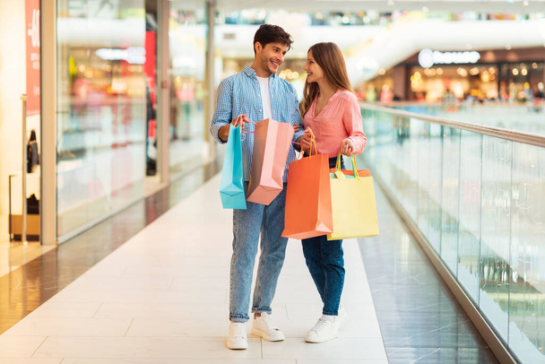 Fröhliches Paar hält Papiereinkaufstaschen in der Hand und kauft gemeinsam neue Kleider, die am Wochenende in einem modernen Einkaufszentrum stehen. Kommerz, großes Vertriebskonzept. Schuss in voller Länge - Foto, Bild
