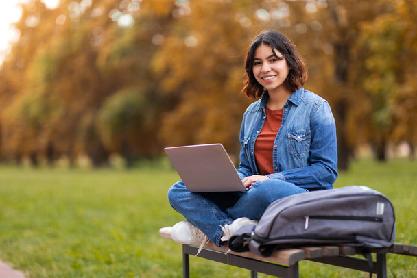 Remote Work Concept. Smiling Young Arab Woman Sitting With Laptop On Bench Outdoors, Portrait Of Cheerful Millennial Middle Eastern Female Freelancer Working With Computer In City Park, Copy Space - Photo, Image