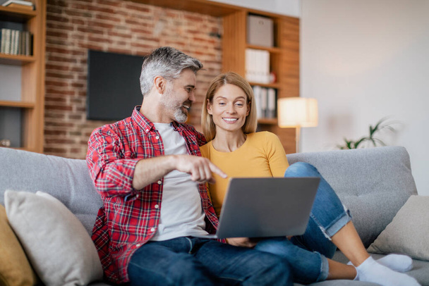 Smiling mature european husband hugs female, pointing finger at computer, sits on sofa in living room interior. Device for social media, chat and internet surfing, great ad and offer due covid-19 - Photo, Image
