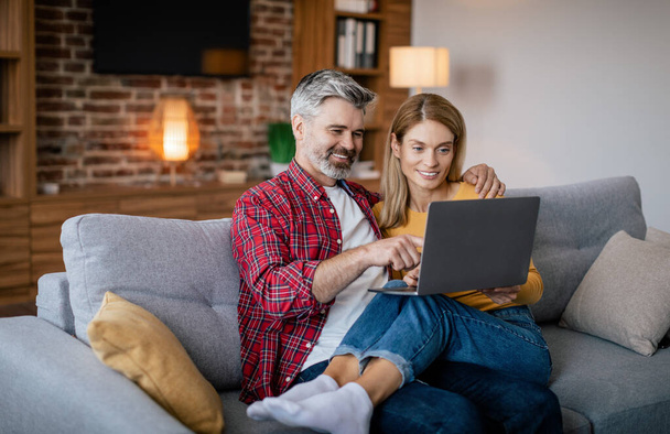 Glad mature european couple hug, look at laptop, surfing in internet, enjoy free time, sit on sofa in living room interior. Video call, meeting remotely, communication, ad and offer during covid-19 - Φωτογραφία, εικόνα