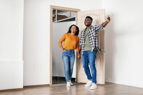 Ενοικιάζεται διαμέρισμα. Ενθουσιασμένοι μαύρο οικογενειακό ζευγάρι Επιλέγοντας και αγοράζοντας νέο σπίτι στέκεται στην ανοιχτή πόρτα του σπιτιού τους. Προσφορά ακινήτων και επιχειρηματική ιδέα. Πλήρες μήκος - Φωτογραφία, εικόνα