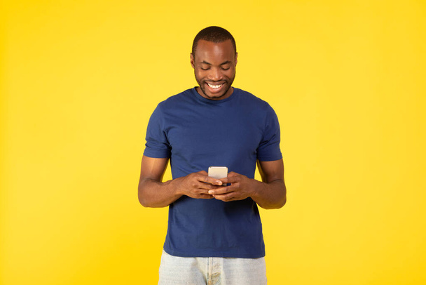 Ευτυχισμένο μαύρο αρσενικό χρησιμοποιώντας το κινητό εφαρμογή στο κινητό τηλέφωνο, κείμενο και Websurfing στέκεται σε κίτρινο φόντο στο στούντιο. Έννοια τεχνολογίας και επικοινωνίας - Φωτογραφία, εικόνα
