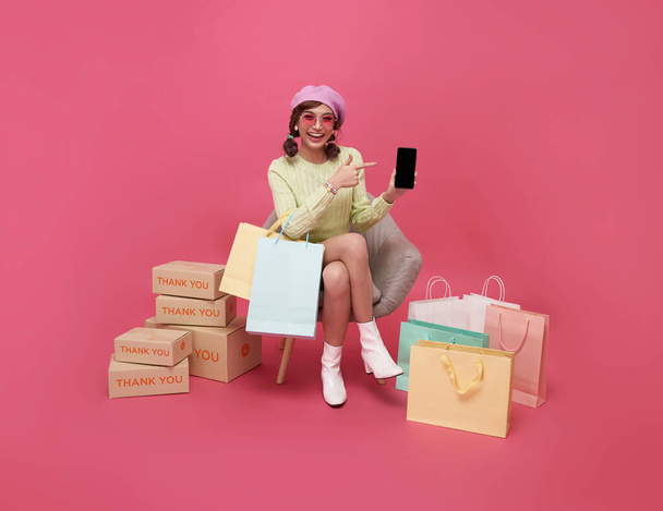 Mujer adolescente asiática feliz sentada en un sofá sosteniendo bolsas de compras y un teléfono inteligente aislado sobre fondo rosa, Shopper o concepto adicto a las compras. - Foto, imagen