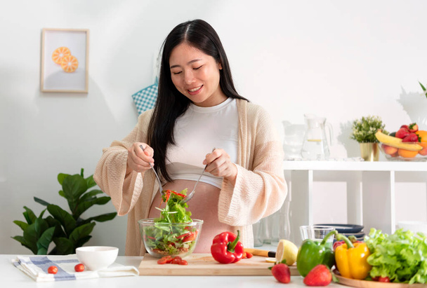 Joyeux asiatique femme enceinte cuisine salade à la maison, faire de la salade verte fraîche, manger de nombreux légumes différents pendant la grossesse, concept de grossesse saine. - Photo, image