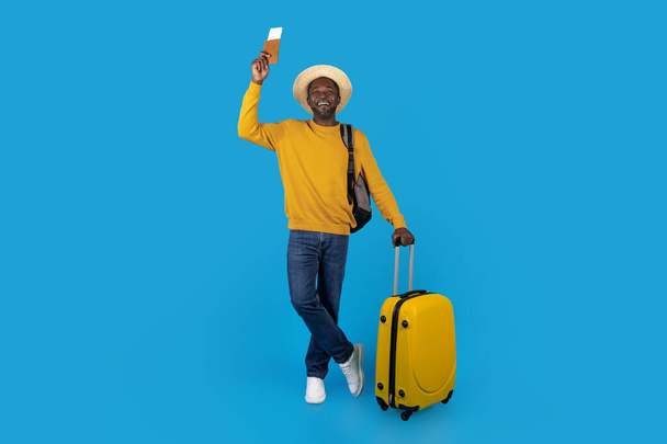 Χαρούμενος Αφροαμερικάνος μεσήλικας με καλοκαιρινό καπέλο τουρίστας που ταξιδεύει μόνος, κουβαλάει κίτρινες αποσκευές, σακίδιο πλάτης, δείχνει διαβατήριο με αεροπορικά εισιτήρια, ποζάρει σε μπλε φόντο στούντιο, αντιγράφει χώρο - Φωτογραφία, εικόνα