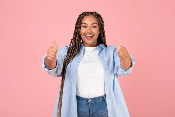 それが好き。陽気な太りすぎアフリカ系アメリカ人女性ピンクの背景の上に立ってカメラに笑顔をポーズアップして親指をジェスチャー,スタジオショット.素晴らしいオファーの広告 - 写真・画像