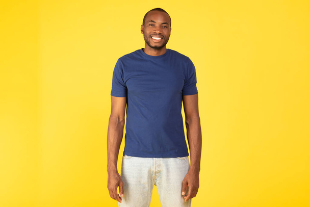 Wesoły czarny facet stojący pozował uśmiechnięty patrząc w kamerę w luźnych ubraniach na żółtym tle. Millennial Man wyrażający pozytywne emocje. Koncepcja męskiej urody. Studio Strzał - Zdjęcie, obraz