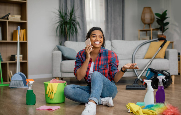 Szczęśliwa młoda czarna kobieta w gumowych rękawiczkach siedzi na podłodze z dodatkami do czyszczenia, dzwoniąc przez telefon we wnętrzu salonu. Odpocznij, zrelaksować się od prac domowych w domu sam, styl życia, plotki i dobre wieści - Zdjęcie, obraz