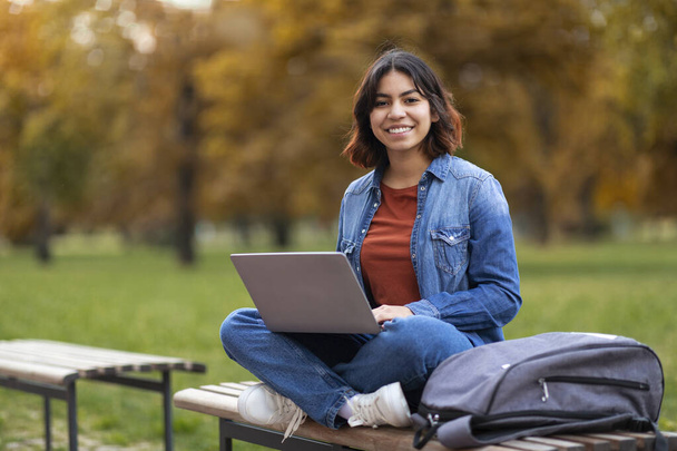 Portrait Of Smiling Young Middle Eastern Female Student With Laptop On Laps Sitting On Bench Outdoors, Happy Millennial Arab Woman With Computer Preparing For Exam Outside In City Park, Copy Space - Foto, Imagem