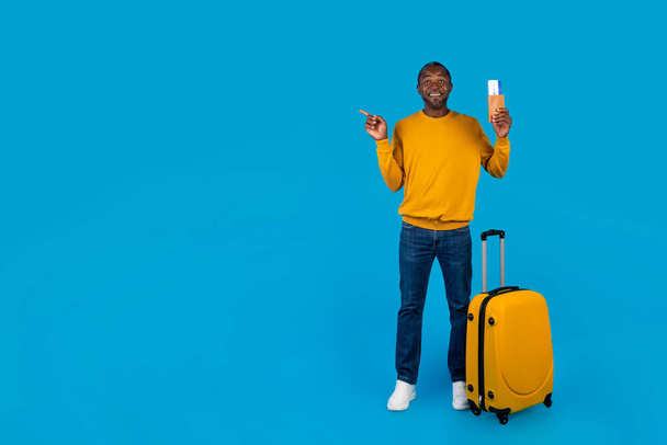 Happy middle aged black man in casual outfit having journey alone, holding passport with flight tickets, standing next to suitcase, pointing at copy space, blue studio background, full length - Photo, Image