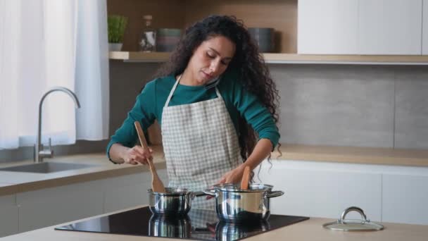 Umtriebige arabische Mutter attraktive lockige Hausfrau Koch trägt Schürze Kochen Frühstück zu Hause Küche Rühren Mahlzeit in Töpfen mit Löffel Zubereitung von Essen für Familie sprechen Mobiltelefon-Handy-Kommunikation - Filmmaterial, Video