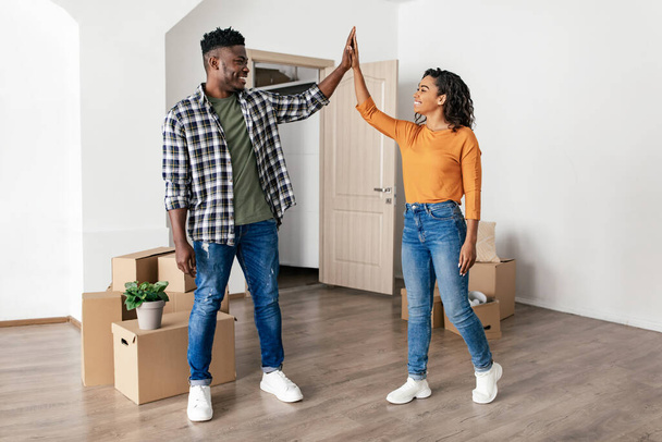 Uusi koti. Iloiset afroamerikkalaiset puolisot antavat yläviittä poseerausta lähellä muuttolaatikoita juhlii uudelleensijoittamista ostamalla uuden talon seisoen sisätiloissa. Kiinteistötarjous - Valokuva, kuva