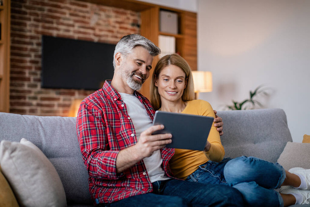 Cheerful mature european couple have video call remotely, chatting by tablet sit on sofa in cozy living room interior. Modern gadget and app for meeting during social distancing, covid-19 pandemic - Foto, Imagen