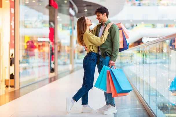 Glückliches Paar beim gemeinsamen Einkaufen mit Einkaufstüten, die am Wochenende in der modernen Mall Indoor stehen. Konsumerismus und großes Verkaufsangebotskonzept. Volle Länge - Foto, Bild