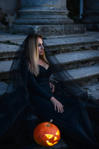 A young woman witch in black clothes sits near the old castle and holds in her hands a luminous pumpkin with a terrible face. - Foto, Imagem
