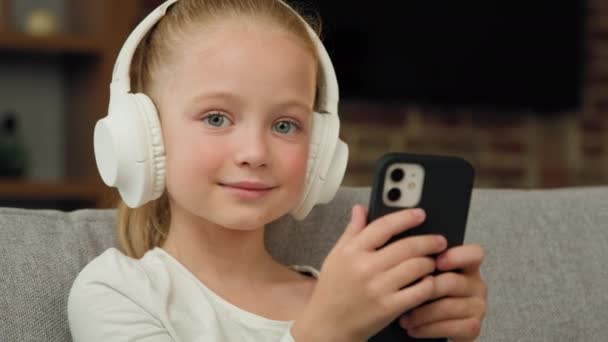 魅力的な白人ブロンドの子供の娘は、モバイル音楽アプリのオーディオプレイリストでヘッドフォンのお気に入りの曲でソファリラックスしたリスニングミュージックに座っている幸せな子供時代を笑顔カメラで音のリズムルックを楽しむ - 映像、動画