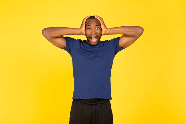 Emocional afroamericano deportista gritando tocando la cabeza mirando a la cámara en shock de pie posando en el fondo del estudio amarillo, usando ropa deportiva. Concepto de Omg - Foto, Imagen