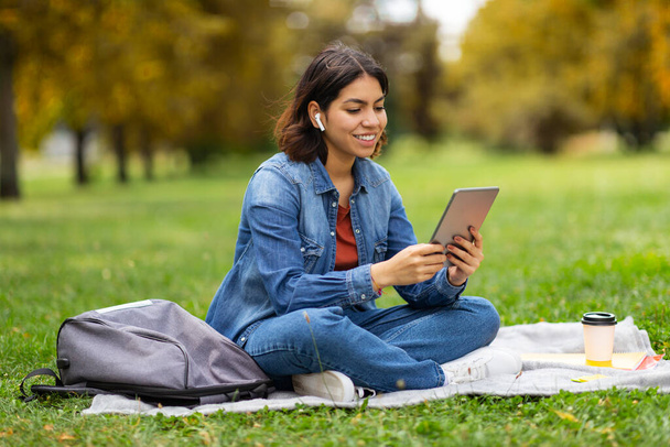 Mujer árabe sonriente joven que relaja en el parque con la tableta digital, hembra milenaria feliz del Medio Oriente que se sienta en el césped al aire libre y que usa el gadget moderno, usando auriculares inalámbricos, espacio de copia - Foto, Imagen