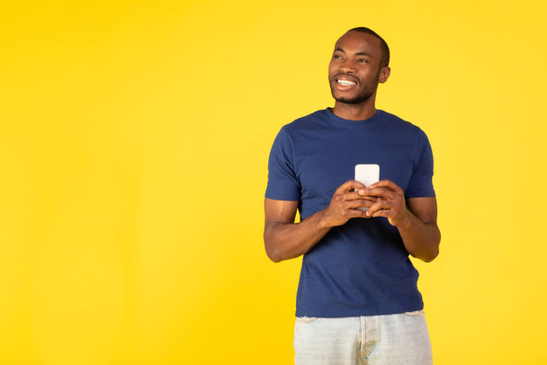 African American Guy Holding Mobile Phone Browsing Internet And Texting Smiling Looking Aside On Yellow Background, Studio Shot. Application Advertisement Concept. Free Space - 写真・画像