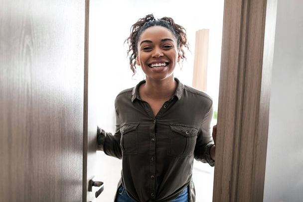 Iloinen musta nainen avaaminen ovi seisoo hymyillen kameralle kotona. Nainen toivottaa sinut tervetulleeksi ja tapaa sinut uudessa asunnossa. Suuri Real Estate Tarjous käsite - Valokuva, kuva