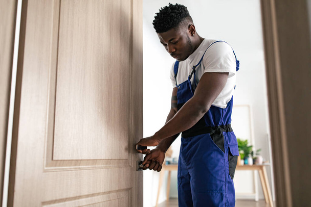 Doors Installation Service. African American Professional Repairman Fixing Door Lock For House Safety Standing Wearing Blue Coverall Uniform At Home. Renovation Concept - Zdjęcie, obraz