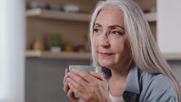 Marzycielska dojrzała kobieta trzymająca filiżankę gorącego napoju rozgrzewające ręce na kubku zapobiegające grypie odwracając wzrok pijąc poranną kawę pozytywnie śniąc przyjemne wspomnienia szczęśliwa starsza babcia relaksująca się w domu - Materiał filmowy, wideo