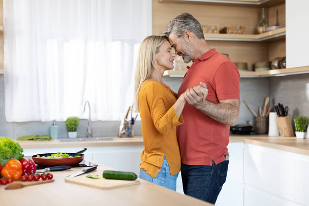 Romantic middle aged couple in casual outfits having fun while cooking together at kitchen, dancing, looking at each other and smiling, happy husband hugging his beautiful wife, copy space - Foto, imagen