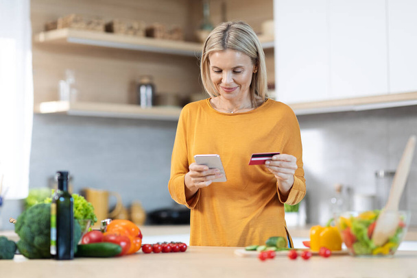Feliz mujer rubia de mediana edad comprando comida en línea, usando el teléfono celular y la tarjeta de crédito de plástico, comprando ingredientes para una cena saludable, cocinando para la familia en casa, espacio para copiar - Foto, imagen