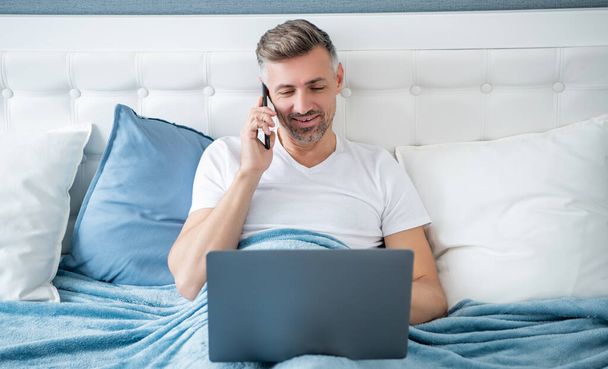 cheerful mature man speaking on phone and working on laptop in bed. - Photo, Image