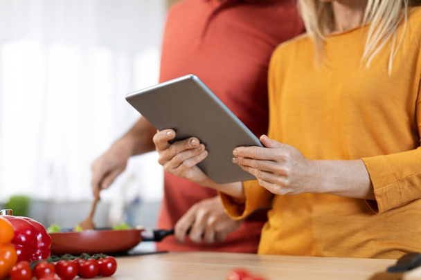 Unrecognizable spouses cooking healthy dinner or lunch together at home, checking recipes online, using digital tablet, reading culinary blog on Internet, cropped, copy space - Фото, изображение