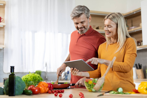 Happy middle aged couple cooking healthy dinner or lunch together at home, checking recipes on Internet, using modern digital tablet, reading culinary blog online, kitchen interior, copy space - 写真・画像