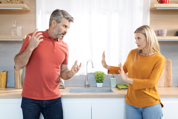Huiselijk geweld, geweld. Emotionele echtgenoten van middelbare leeftijd hebben ruzie in de keuken, woedende blonde vrouw en grijsharige man schreeuwen tegen elkaar, ervaren relatiecrisis, zijaanzicht - Foto, afbeelding