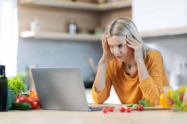 Zmęczona blondynka samotna matka patrząca na nowoczesny ekran laptopa i dotykająca głowy podczas gotowania dla rodziny w domu, cierpiąca na migrenę, wnętrze kuchni, przestrzeń do kopiowania - Zdjęcie, obraz