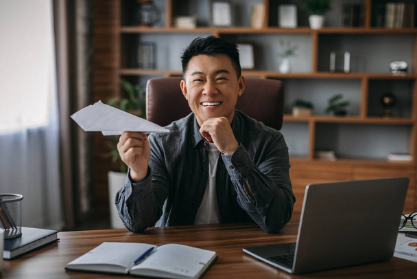 Happy adult chinese man rest from work, make paper airplane in office interior. Take break, successful home business, new normal. Entrepreneur or investor, develop, planning and financial strategies - 写真・画像