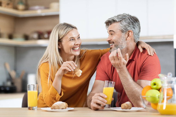 Cheerful loving middle aged man and woman sitting at kitchen table, having snack, talking at home, married couple enjoying time together, cuddling, talking, eating pastry, drinking juice, copy space - Fotoğraf, Görsel