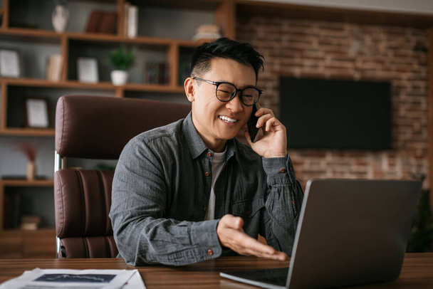 Cheerful mature japanese man in glasses calls by phone, works with laptop at table in office interior. Businessman talking with client, professional investor and new project during social distance - Foto, immagini
