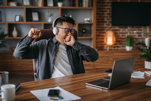 Уставший взрослый корейский менеджер в очках и наушниках зевает и отдыхает от работы в домашнем офисе. Развитие, деловая и социальная дистанцированность, перерыв, дедлайн и поддержка клиентов - Фото, изображение