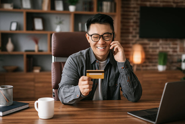 Glad middle aged korean man in glasses calls by phone use laptop and credit card, shopping online in office interior. Professional working new start up, credit and finance, businessman enjoy sale - 写真・画像