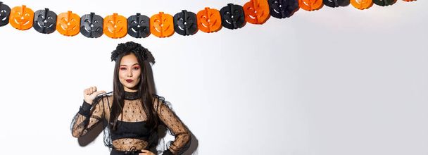 Itsevarma aasialainen nainen tyylikäs goottilainen mekko ja musta seppele osoittaa itseään kuin ammattilainen, seisoo halloween koristeet. - Valokuva, kuva