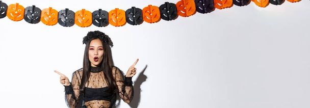 Radostná asijská žena v gotických krajkových šatech slaví Halloween, ukazuje prsty bokem, stojí proti bílému pozadí s dýňovými stranickými stuhami. - Fotografie, Obrázek