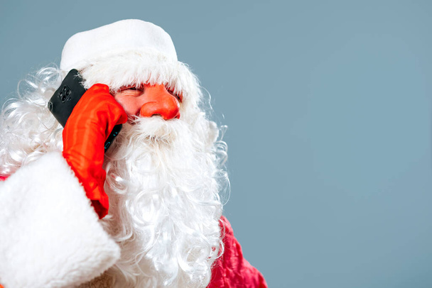Santa Claus on blue studio background in red hat with white beard holds a smartphone with his hand to his ear and speaks on it. An interesting conversation about gifts for children for the new year. - Foto, Bild