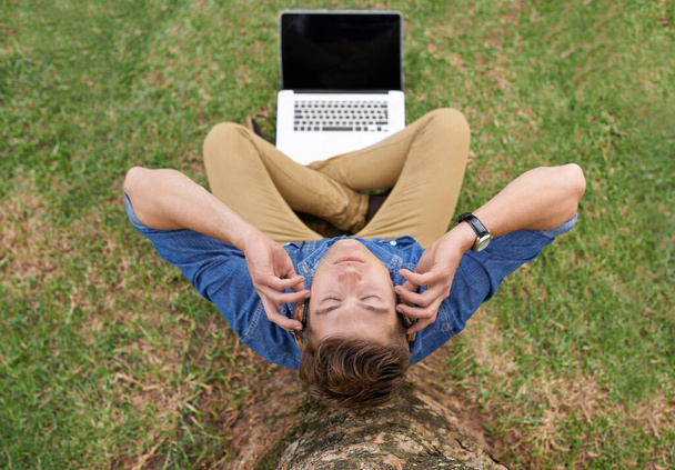 Alejarse de todo con música. Foto de ángulo alto de un joven sentado frente a una computadora portátil afuera y escuchando música - Foto, Imagen